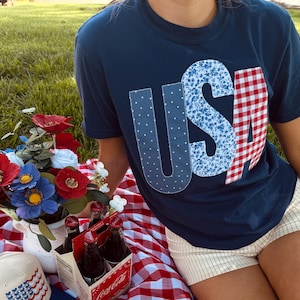 May include: A navy blue t-shirt with the letters USA appliqued in polka dot, floral, and red gingham patterns. A cream baseball cap with a stylized American flag embroidery, a bouquet of red and blue flowers, and a four-pack of Coca-Cola glass bottles.