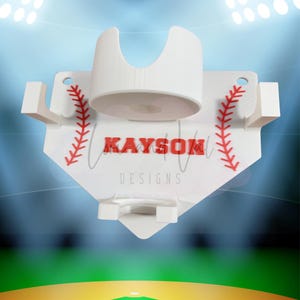 May include: White baseball-themed wall mount with red stitching details and the text "KAYSON DESIGNS". The mount features a cup holder and two side hooks. The background shows a baseball field under stadium lights.