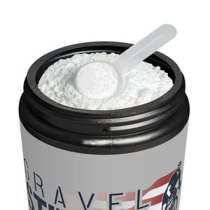 May include: A cylindrical container filled with white powder, topped with a black rim and a clear plastic scoop. The container has a gray label with the words "GRAVEL" and "STRENGTH" and a graphic of a cyclist and the American flag.