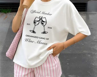 Può includere: T-shirt bianco con la scritta "Official Member EST. 2026 ORGANIZED GANG OF Wine Moms" e una grafica di due calici di vino che si toccano. La persona indossa pantaloncini a righe rosa e bianche.
