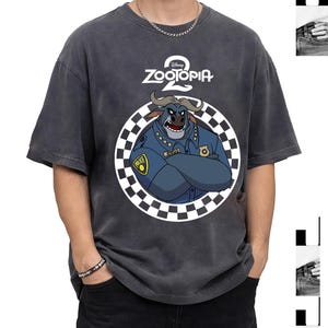 May include: Dark grey Zootopia 2 t-shirt with a cartoon bull character in a police uniform inside a chequered circle. The Disney logo is above the character. The shirt has short sleeves and a relaxed fit.