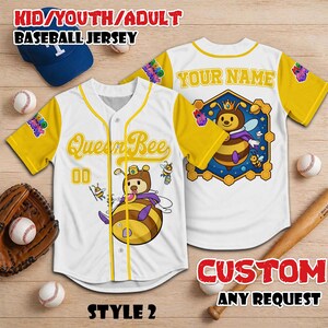 Puede incluir: Camiseta de b&eacute;isbol blanca con mangas y ribetes amarillos. La parte delantera presenta una abeja de dibujos animados con el texto "Queen Bee" y el n&uacute;mero "00". La parte trasera tiene un gr&aacute;fico de abeja y espacio para un nombre. Ropa tem&aacute;tica de b&eacute;isbol.