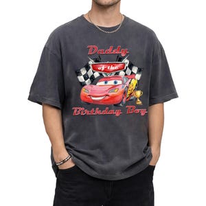 May include: Dark gray t-shirt featuring a graphic of Lightning McQueen from the movie Cars, with the text "Daddy of the Birthday Boy." The design includes a checkered flag and a trophy.