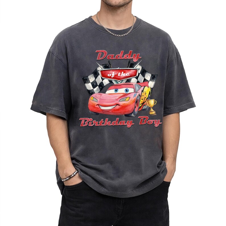 May include: Dark gray t-shirt featuring a graphic of Lightning McQueen from the movie Cars, with the text "Daddy of the Birthday Boy." The design includes a checkered flag and a trophy.