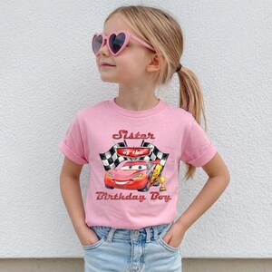 May include: A pink t-shirt featuring a graphic of a red race car with the text "Sister of the Birthday Boy." The design includes checkered flags and a golden trophy. The shirt is worn by a young girl wearing pink heart-shaped sunglasses.
