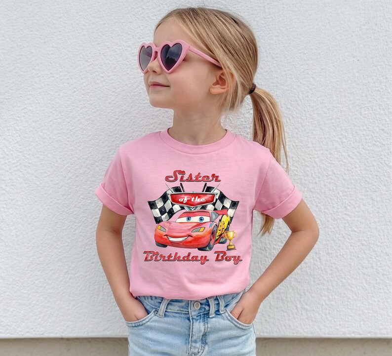 May include: A pink t-shirt featuring a graphic of a red race car with the text "Sister of the Birthday Boy." The design includes checkered flags and a golden trophy. The shirt is worn by a young girl wearing pink heart-shaped sunglasses.