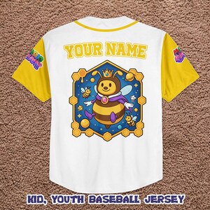 Puede incluir: Camiseta de b&eacute;isbol blanca para ni&ntilde;os con mangas y cuello amarillos. La camiseta presenta un personaje de abeja de dibujos animados con una corona y una capa dentro de un marco hexagonal. El texto "YOUR NAME" est&aacute; encima del gr&aacute;fico y "KID, YOUTH BASEBALL JERSEY" debajo.