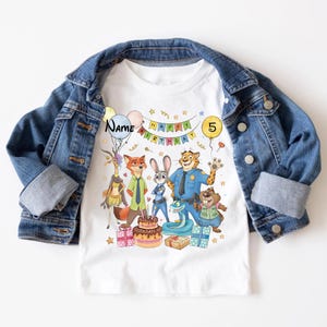 K&ouml;nnte beinhalten: Wei&szlig;es T-Shirt mit einem farbenfrohen Zootopia-Motiv zum Geburtstag. Das Design zeigt Charaktere, einen Kuchen, Geschenke und die Texte "Happy Birthday" und "5". Das T-Shirt wird unter einer blauen Jeansjacke getragen.