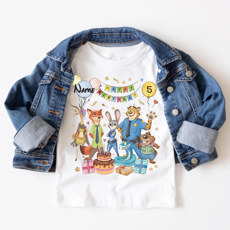 K&ouml;nnte beinhalten: Wei&szlig;es T-Shirt mit einem farbenfrohen Zootopia-Motiv zum Geburtstag. Das Design zeigt Charaktere, einen Kuchen, Geschenke und die Texte "Happy Birthday" und "5". Das T-Shirt wird unter einer blauen Jeansjacke getragen.