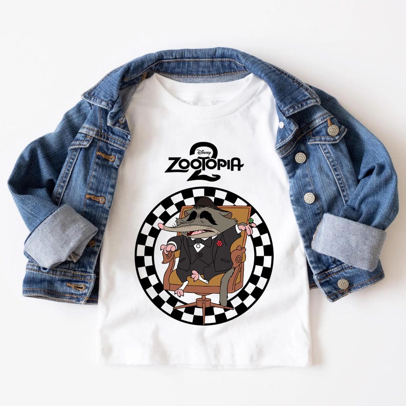 May include: White t-shirt with a cartoon illustration of a Zootopia character in a suit, seated in a chair within a black and white checkered circle. The shirt is overlaid by a denim jacket.