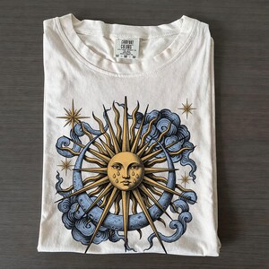 May include: A cream-colored short-sleeve t-shirt featuring a graphic print of a golden sun with a face and teardrops, surrounded by a blue crescent moon, swirling blue clouds, and gold stars. The inner neck label reads Comfort Colors M M.