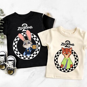 May include: Two children's t-shirts featuring characters from the Disney film Zootropolis. One black shirt displays Judy Hopps, the bunny police officer, and the other is cream-coloured with Nick Wilde, the fox. Both shirts have the Zootropolis logo.
