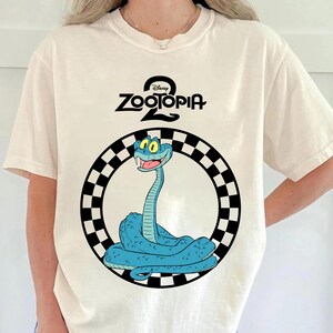 May include: Cream-coloured t-shirt with a cartoon image of a blue snake inside a black and white chequered circle. The text "Disney Zootopia 2" is above the snake illustration.