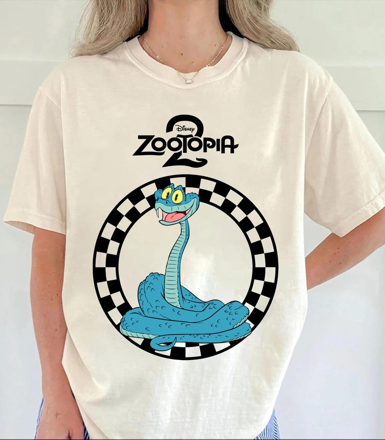 May include: Cream-coloured t-shirt with a cartoon image of a blue snake inside a black and white chequered circle. The text "Disney Zootopia 2" is above the snake illustration.