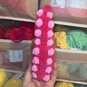 May include: A pink crocheted octopus with large pink pom-poms. The octopus is being held in a hand.