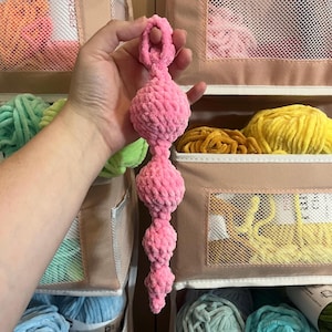 May include: A pink crocheted ball chain with a loop at the top. The chain is made of multiple pink balls connected together. The chain is being held by a hand.