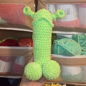 May include: A handmade, light green crochet sculpture of a phallus with two testicles and two horn-like protrusions. The sculpture is made of a soft, textured yarn. The background includes yarn and storage containers.