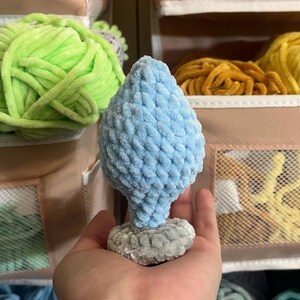 May include: A light blue crocheted tree ornament with a grey base. The ornament is sitting on a hand.