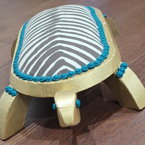 May include: A gold and turquoise zebra-striped footstool shaped like a turtle. The footstool has a gold base and turquoise accents.