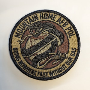 May include: A brown and tan embroidered patch with a black snake wrapped around a globe. The text reads "Mountain Home AFB POL Going Nowhere Fast Without Our Gas".