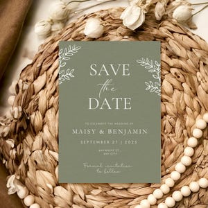 Könnte beinhalten: Eine salvengrüne Hochzeitseinladung mit weißem Text und floralen Akzenten. Der Text lautet "Save the Date" und enthält die Namen Maisy & Benjamin, das Datum 27. September 2025 und eine Adresse. Die Einladung befindet sich auf einem gewebten Hintergrund.