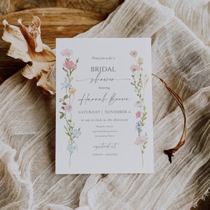 May include: A white invitation card for a bridal shower, featuring floral illustrations in pink, blue, and yellow. The text reads "BRIDAL shower honoring Hannah Brown" with event details. The card is on a textured, neutral-toned surface.