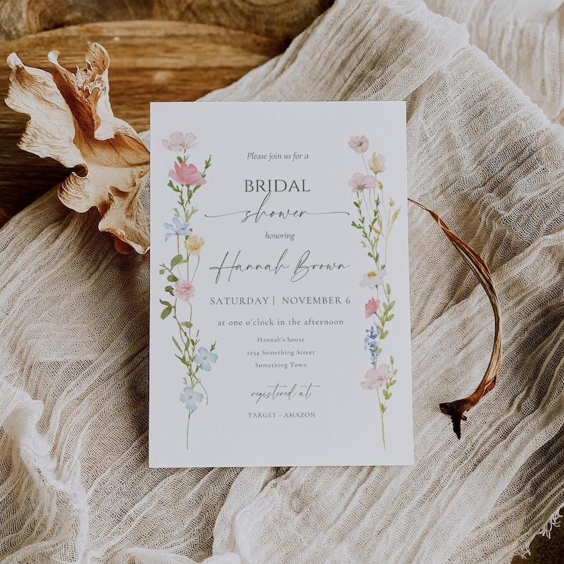May include: A white invitation card for a bridal shower, featuring floral illustrations in pink, blue, and yellow. The text reads "BRIDAL shower honoring Hannah Brown" with event details. The card is on a textured, neutral-toned surface.
