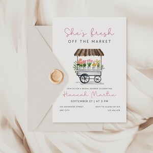 Peut inclure: Une invitation de f&ecirc;te pr&eacute;nuptiale blanche avec une illustration aquarelle d'un chariot &agrave; fleurs. Le texte indique "She's fresh off the market" et comprend le nom de la mari&eacute;e, Hannah Martin, la date et l'heure de la f&ecirc;te, ainsi que les informations RSVP.