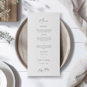 May include: A minimalist wedding menu card with a mountain range illustration at the bottom. The menu features a three-course meal: Creamy Butternut Squash Soup, Beef Tenderloin with a Red Wine Reduction, and Seared Scallops with a Lemon Beurre Blanc. The card is printed on white paper with black text and is set on a wooden table.