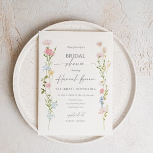 May include: A bridal shower invitation featuring floral accents in pink, blue, and yellow. The invitation includes the text "Please join us for a BRIDAL shower honoring Hannah Brown" and event details. The invitation is on a white plate.