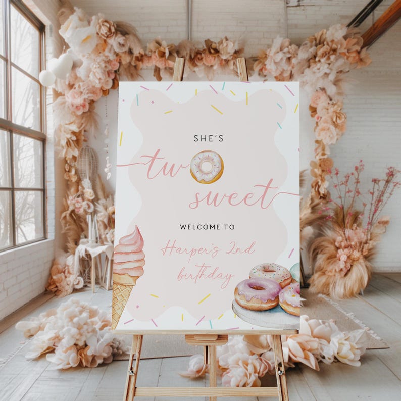 May include: A pink sign on a wooden easel, featuring the text "She's Two Sweet" and "Welcome to Harper's 2nd birthday". The sign includes illustrations of a donut and an ice cream cone.