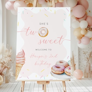 May include: A pink and white welcome sign on an easel, with the text "She's Two Sweet" and "Welcome to Harper's 2nd birthday." The sign features illustrations of donuts and an ice cream cone, with a balloon arch in the background.