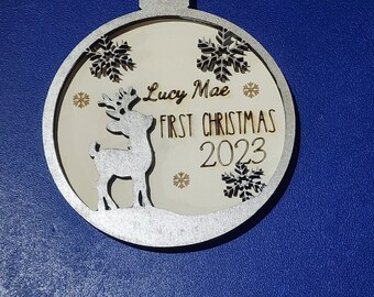 Op de afbeelding: Een zilveren en houten kerstbal met de tekst "Lucy Mae First Christmas 2023". De ornamenten hebben een rendier silhouet, sneeuwvlokken en een cirkelvormig ontwerp. De achtergrond is lichtcrème van kleur.