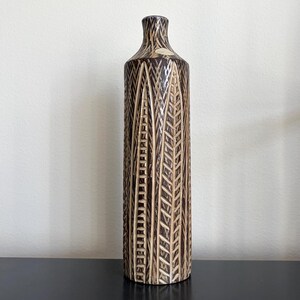May include: A tall, cylindrical ceramic vase featuring a dark brown glaze with a carved, light beige leaf-like pattern running vertically along the entire body. The vase has a narrow neck and a small, circular opening at the top.