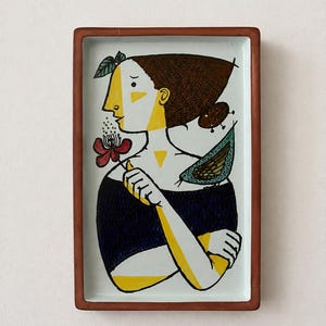 Puede incluir: Una bandeja rectangular de cerámica con borde marrón que presenta una ilustración estilizada de una persona con cabello castaño, una blusa azul oscuro y detalles amarillos. La persona sostiene una flor roja y un pequeño pájaro verde azulado descansa sobre su hombro.