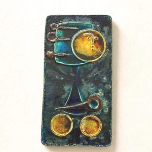 Puede incluir: Una placa de pared rectangular de cerámica con un esmalte moteado en azul profundo y verde azulado. La superficie muestra una composición abstracta con una cara circular amarilla, tres pequeños bucles de metal, una forma de lágrima oscura y dos círculos amarillos.