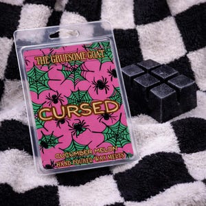 May include: A clear plastic package containing a pink card with a spider web and spider design. The text on the card reads "THE GRUESOME GOAT", "CURSED", and "CUCUMBER MELON HAND-POURED WAX MELTS". Next to the package is a black wax melt.