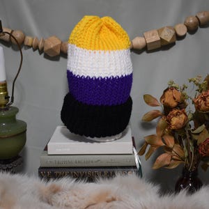 May include: A handmade, knitted hat in the non-binary pride flag colors: yellow, white, purple, and black. The hat is displayed on a stack of books, with a vintage lamp and dried flowers in the background.
