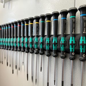 May include: A set of 25 black and green screwdrivers with different sized tips, hanging on a wall-mounted holder. The holder has a green and black design with the letter "H" repeated on it.