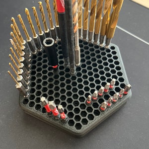 May include: A black hexagonal drill bit holder with a honeycomb design. The holder is filled with various drill bits, including gold-colored drill bits and red-tipped screwdriver bits.