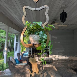 May include: A wooden cat-shaped hanging planter with a heart-shaped accent. The planter holds a green leafy plant in a white pot. A wooden cat silhouette is on the ground. The scene is set on a porch with wicker furniture.