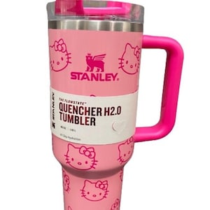 Pode incluir: Caneca Stanley Quencher H2.0 rosa com alça e tampa rosa. A caneca apresenta um padrão repetido de rostos Hello Kitty. O texto "STANLEY" e "QUENCHER H2.0 TUMBLER" são visíveis.