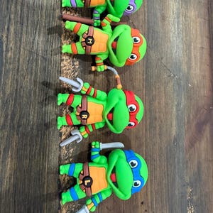 Puede incluir: Cuatro dijes de zapatos de las Tortugas Ninja. Cada tortuga es verde con una máscara de diferente color: morado, naranja, rojo y azul. Cada una tiene un arma diferente y las letras D, M, R y L en sus cinturones.