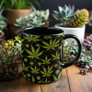 May include: A black ceramic mug with a green marijuana leaf pattern. The mug is sitting on a wooden table with green plants in the background.