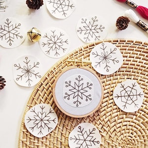 May include: A wooden embroidery hoop containing light grey fabric with a stitched dark brown snowflake design, surrounded by several circular fabric patches featuring various dark brown snowflake patterns, pinecones, and embroidery thread.