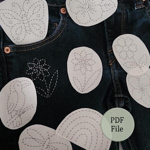Può includere: Jeans in denim blu scuro con diversi modelli in carta bianca con motivi da ricamo a linee tratteggiate, tra cui una farfalla, fiori, un uccello, una libellula e un arcobaleno. Un'etichetta circolare verde chiaro riporta il testo PDF File.