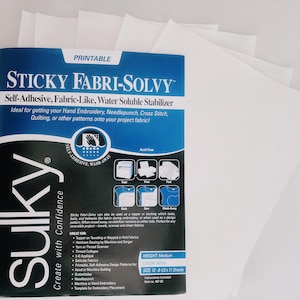 Puede incluir: Un paquete de hojas de estabilizador soluble en agua, autoadhesivas, tipo tela "STICKY FABRI-SOLVY". El paquete es azul y blanco, con texto que indica que es imprimible e ideal para bordado a mano, needlepunch, punto de cruz y acolchado. Las hojas son blancas.