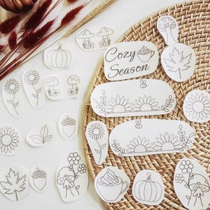 May include: A collection of white paper stickers featuring black line art illustrations of pumpkins, sunflowers, mushrooms, acorns, maple leaves, and cups of coffee with the text Cozy Season, arranged on a round woven rattan surface.