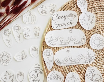 May include: A collection of white paper stickers featuring black line art illustrations of pumpkins, sunflowers, mushrooms, acorns, maple leaves, and cups of coffee with the text Cozy Season, arranged on a round woven rattan surface.