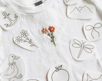Puede incluir: Camiseta blanca con un pequeño diseño floral bordado y varios parches termoadhesivos con varios dibujos lineales. Los parches incluyen una ancla, flores, un pato, un lazo, una libélula, una cordillera y un arcoíris.
